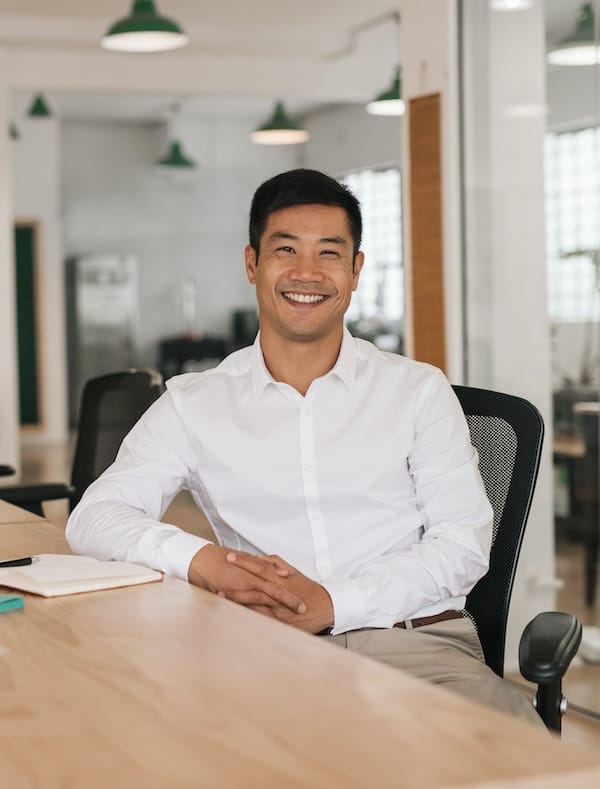 Portrait of a young Asian businessman smiling while using a laptop and writing down notes while sitting at his desk in a modern office